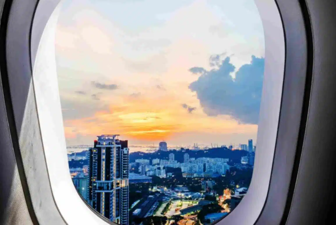 Singapore city skyline at sunset viewed through an airplane window, showing tall buildings, warm sky colors, and a distant coastline.