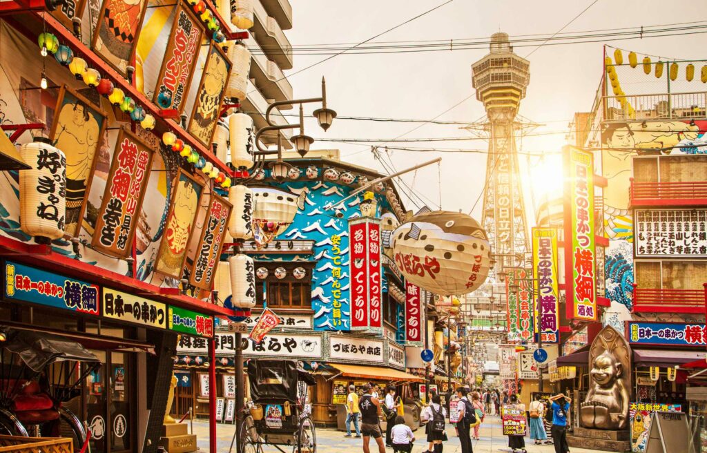Colorful street in Osaka, Japan, filled with lanterns, vibrant signs, and Tsutenkaku Tower in the background at sunset.
