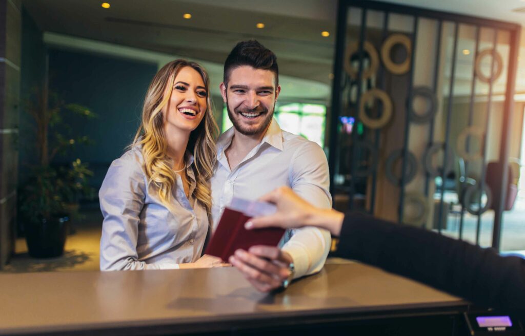 Smiling couple at a hotel reception desk receiving a passport from the receptionist.