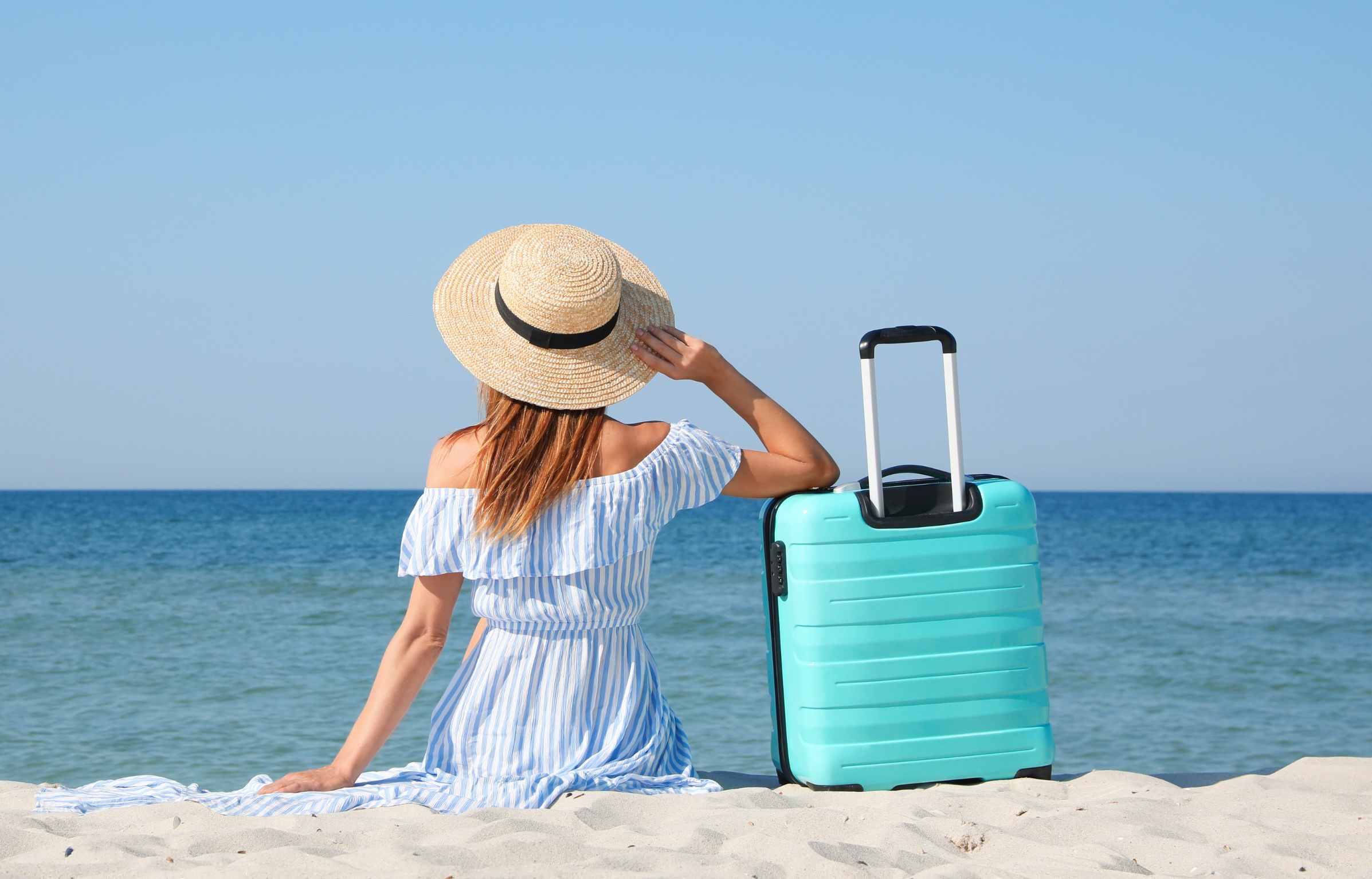 Woman with suitcase sitting on sandy beach near sea, back view