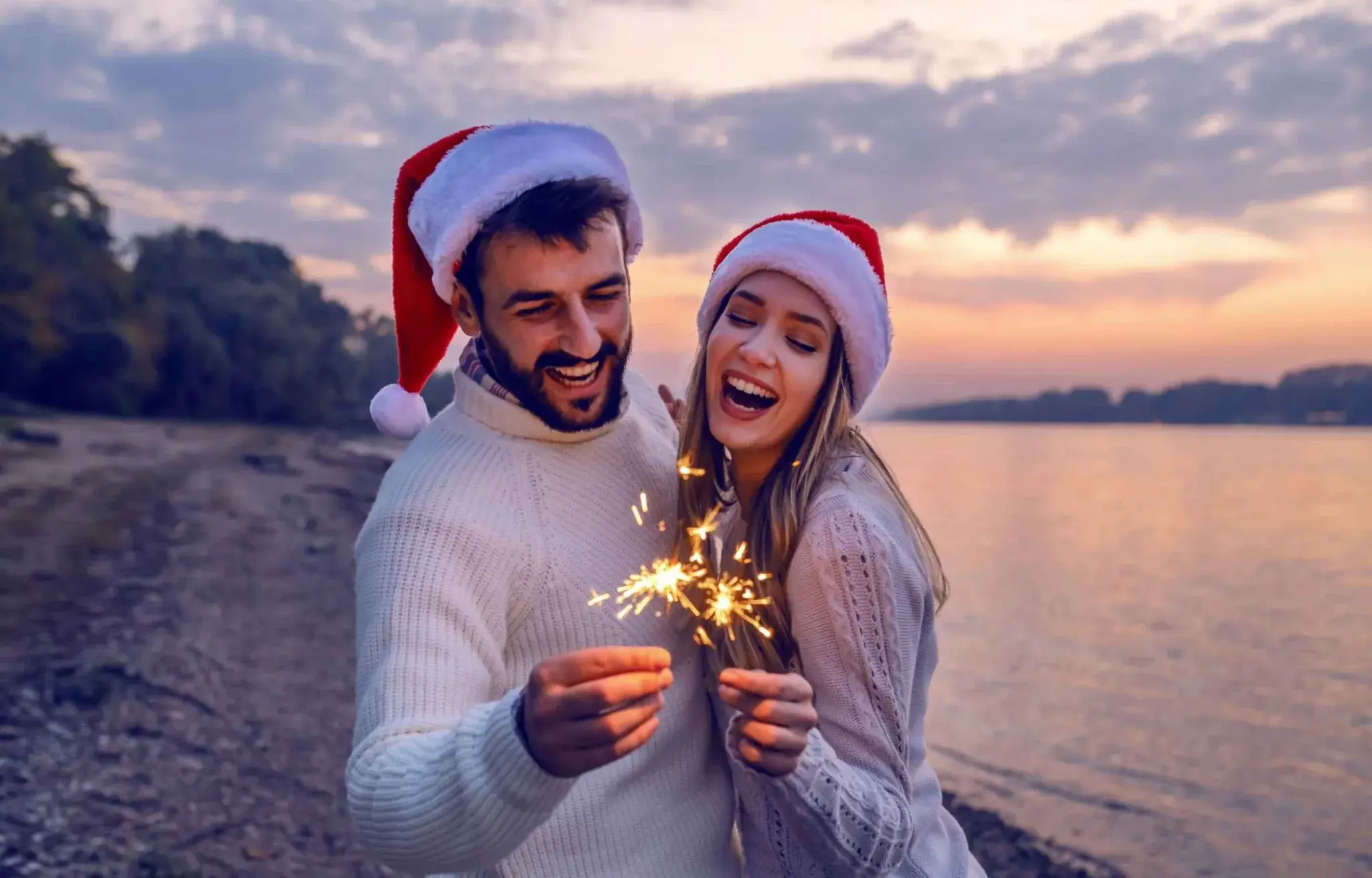 couple wearing santa hats holding sparklers by a lakeside at sunset, celebrating a festive Christmas moment outdoors