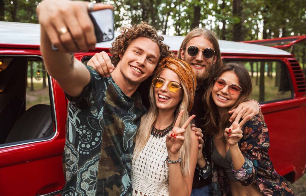 Group of young hippy men and women smiling, and taking selfie on mobile phone near vintage minivan into the nature