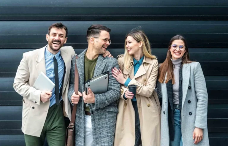 Group of young professionals standing together outdoors, smiling and holding work bags and devices.