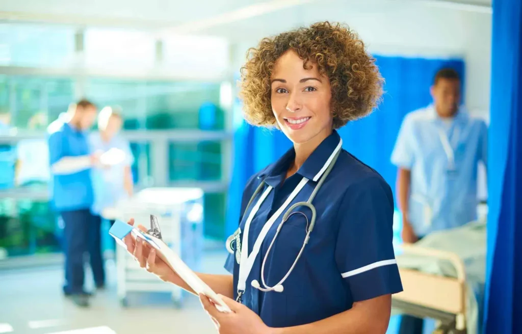 Smiling nurse holding a clipboard in a hospital ward with patients and staff in the background.