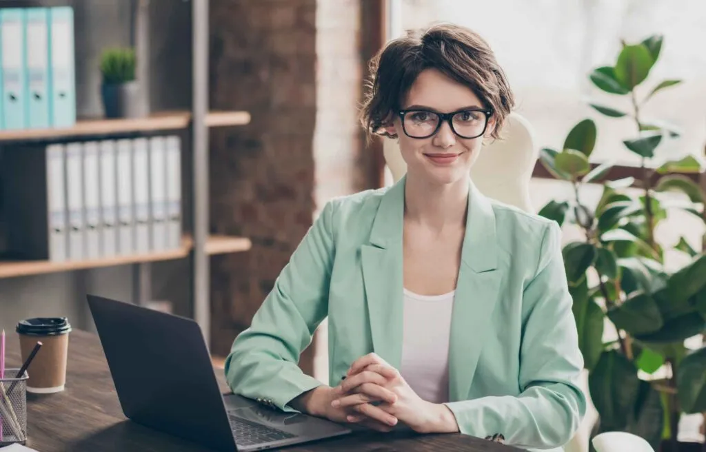 Professional woman in glasses sitting at desk with laptop in a modern office.