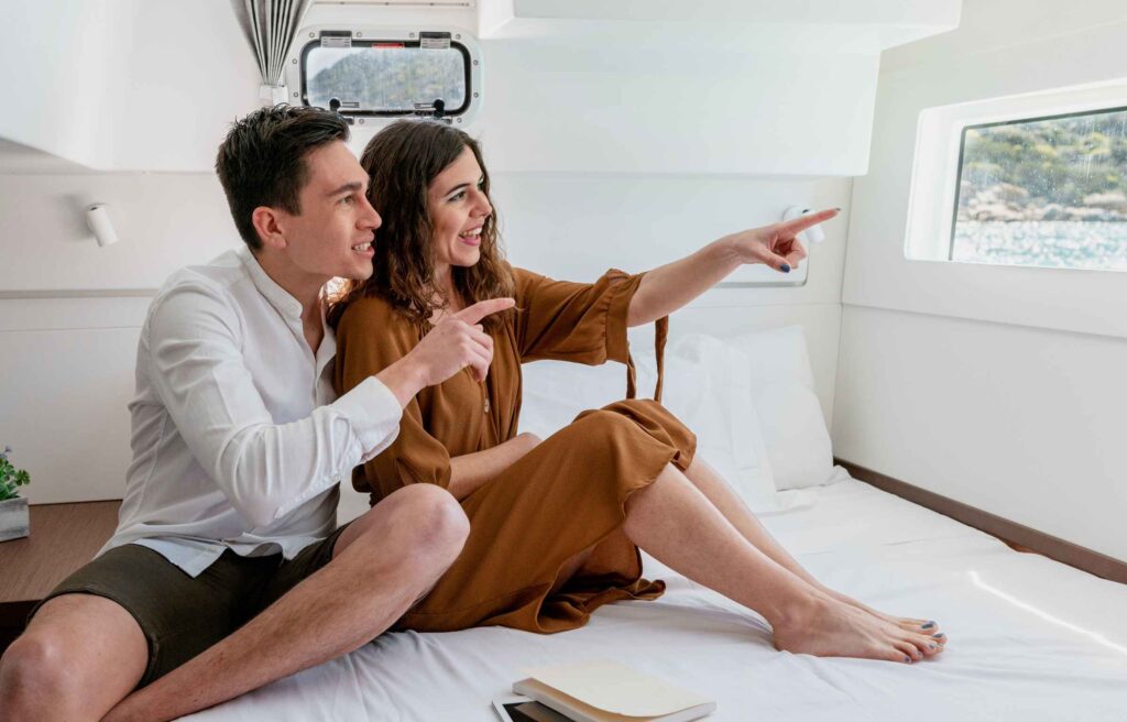 Side view of cheerful loving couple sitting on bed in cabin and looking out of window while sailing on catamaran during vacation