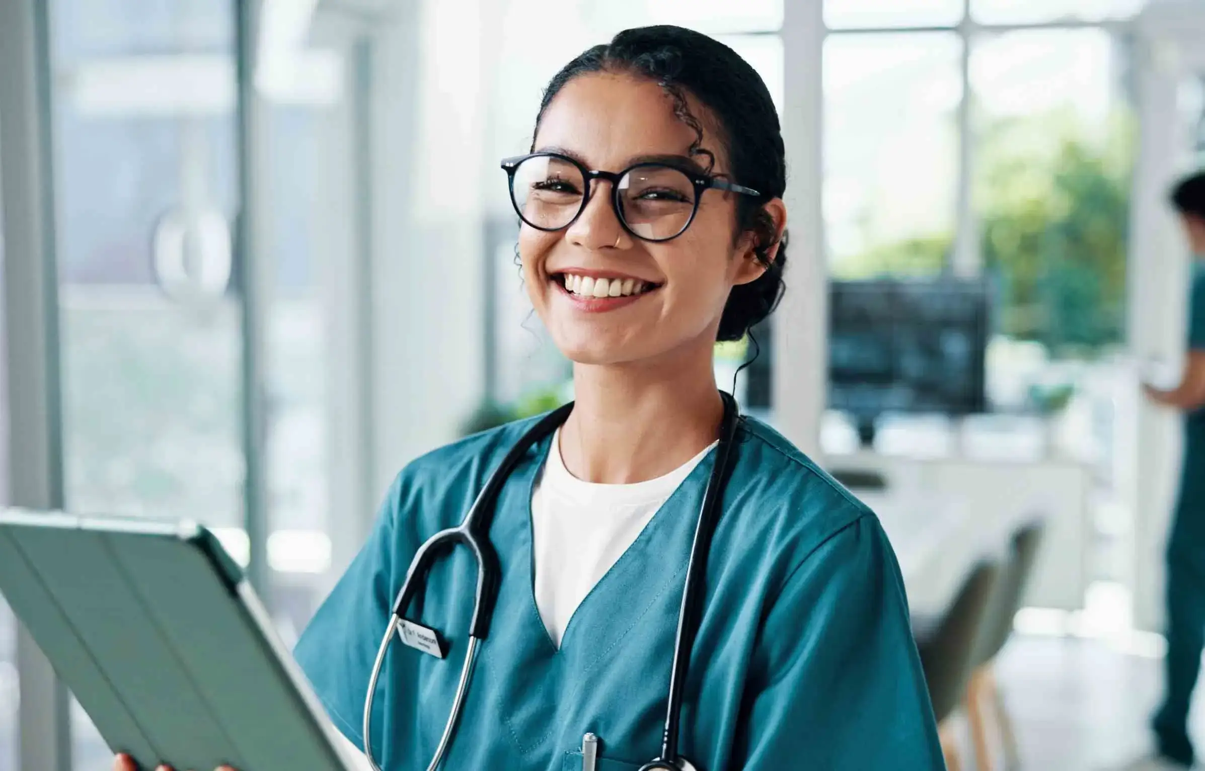 Smiling nurse wearing glasses holding a tablet inside a modern hospital environment.