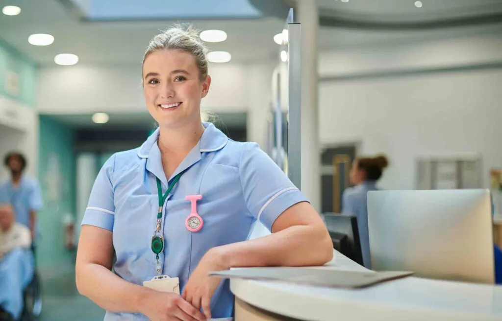Hospital nurse standing at a reception desk with a laptop and patient area behind her.