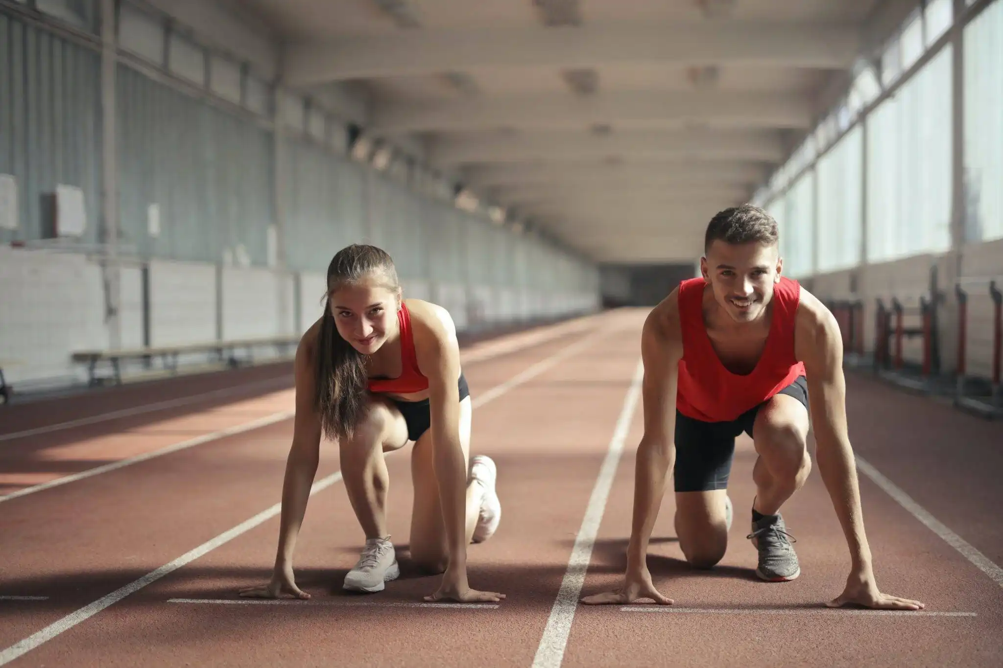 Two athletes crouched at the starting line on an indoor track, ready to sprint.