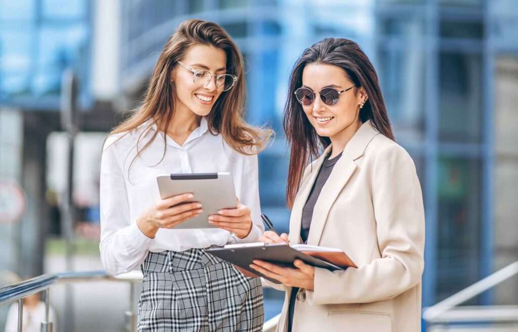 Two young business lady with tablet and notebook walking outdoors near modern business center