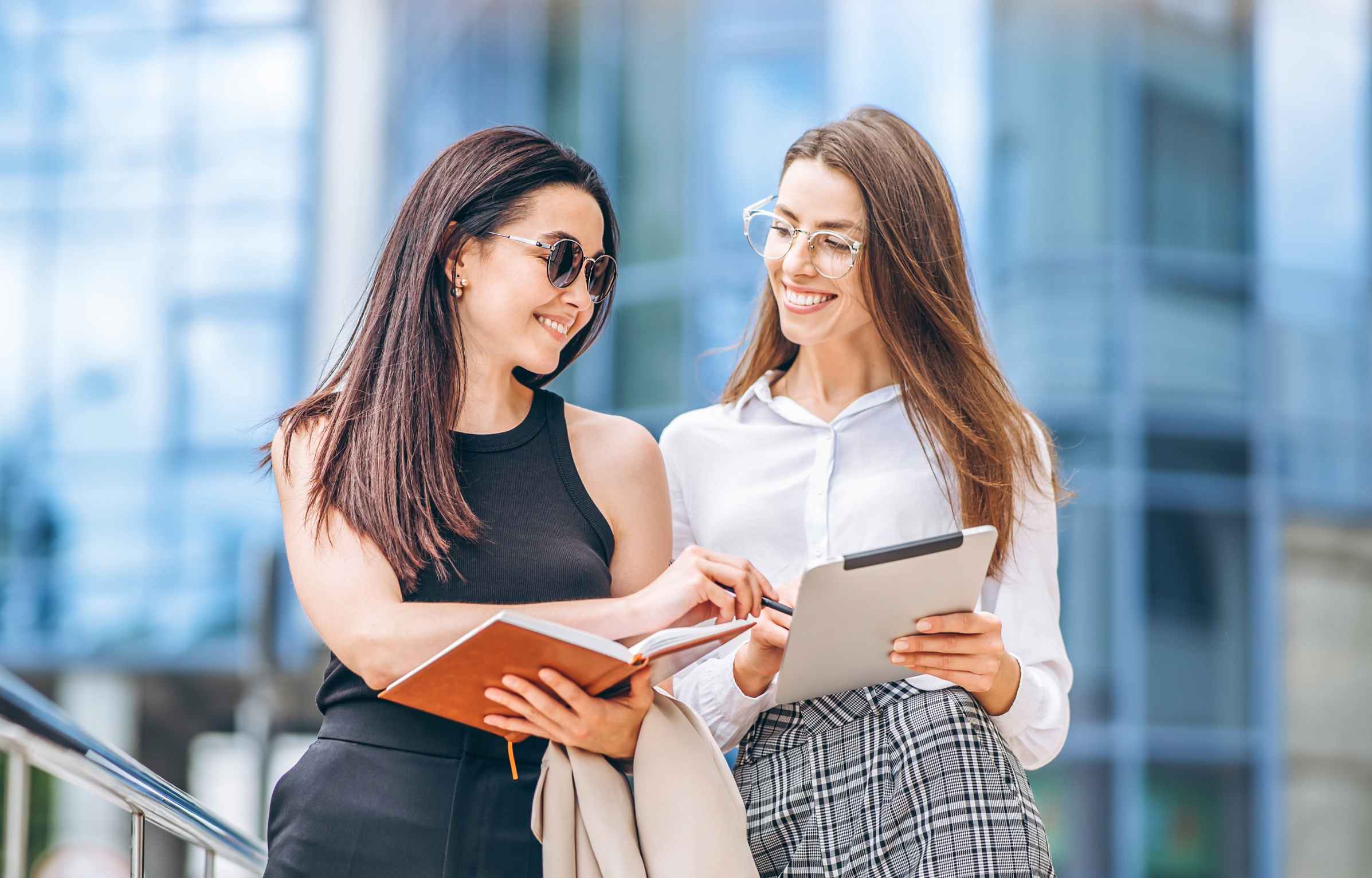 Two young business lady with tablet and notebook walking outdoors near modern business center