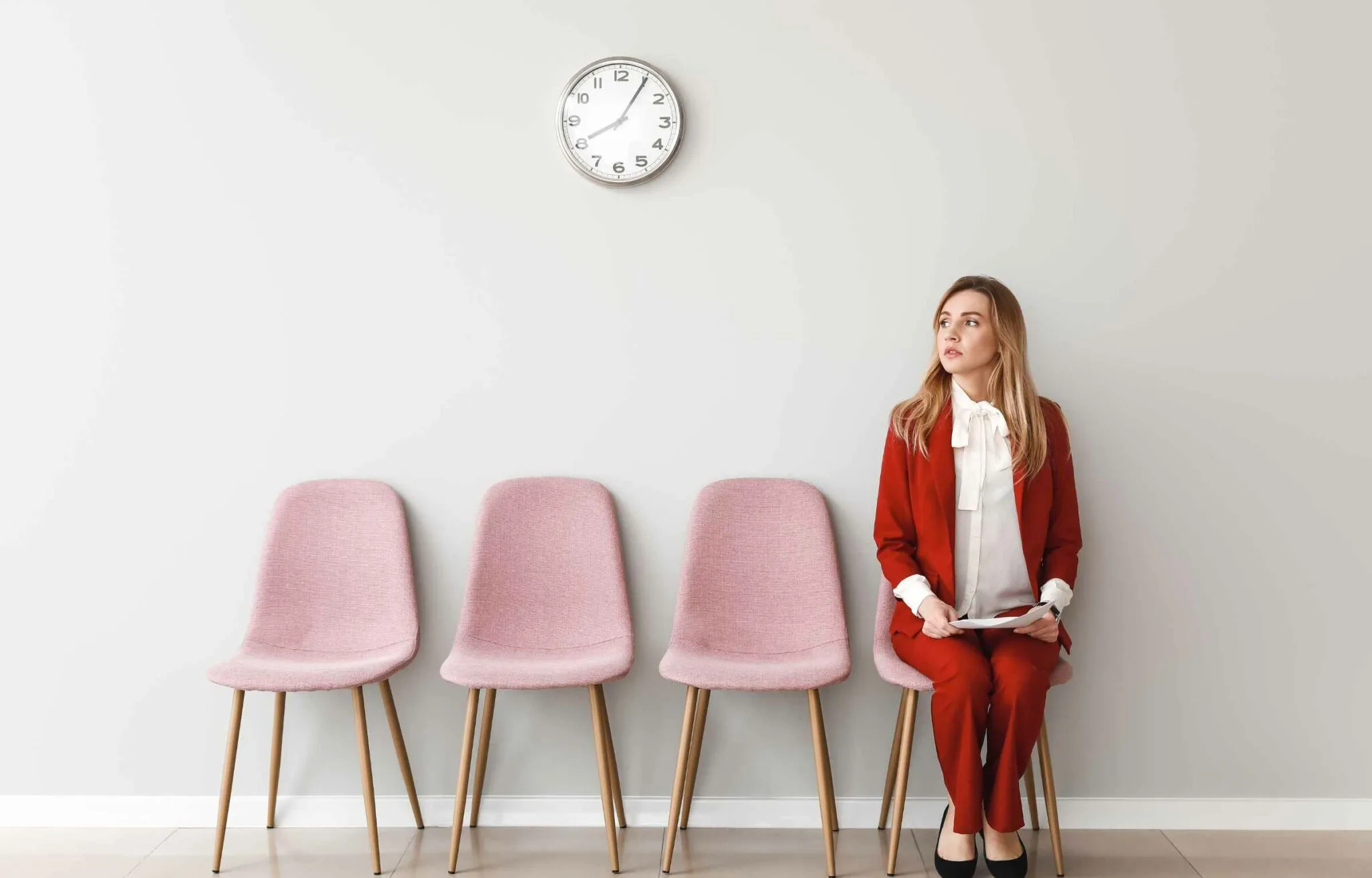 Woman in formal attire sitting on a chair in a waiting area, holding documents and waiting for an interview.