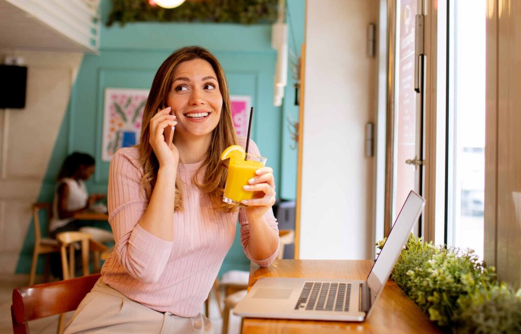 Young woman using mobile phone while working on laptop and drinking fresh orange juice in the cafe