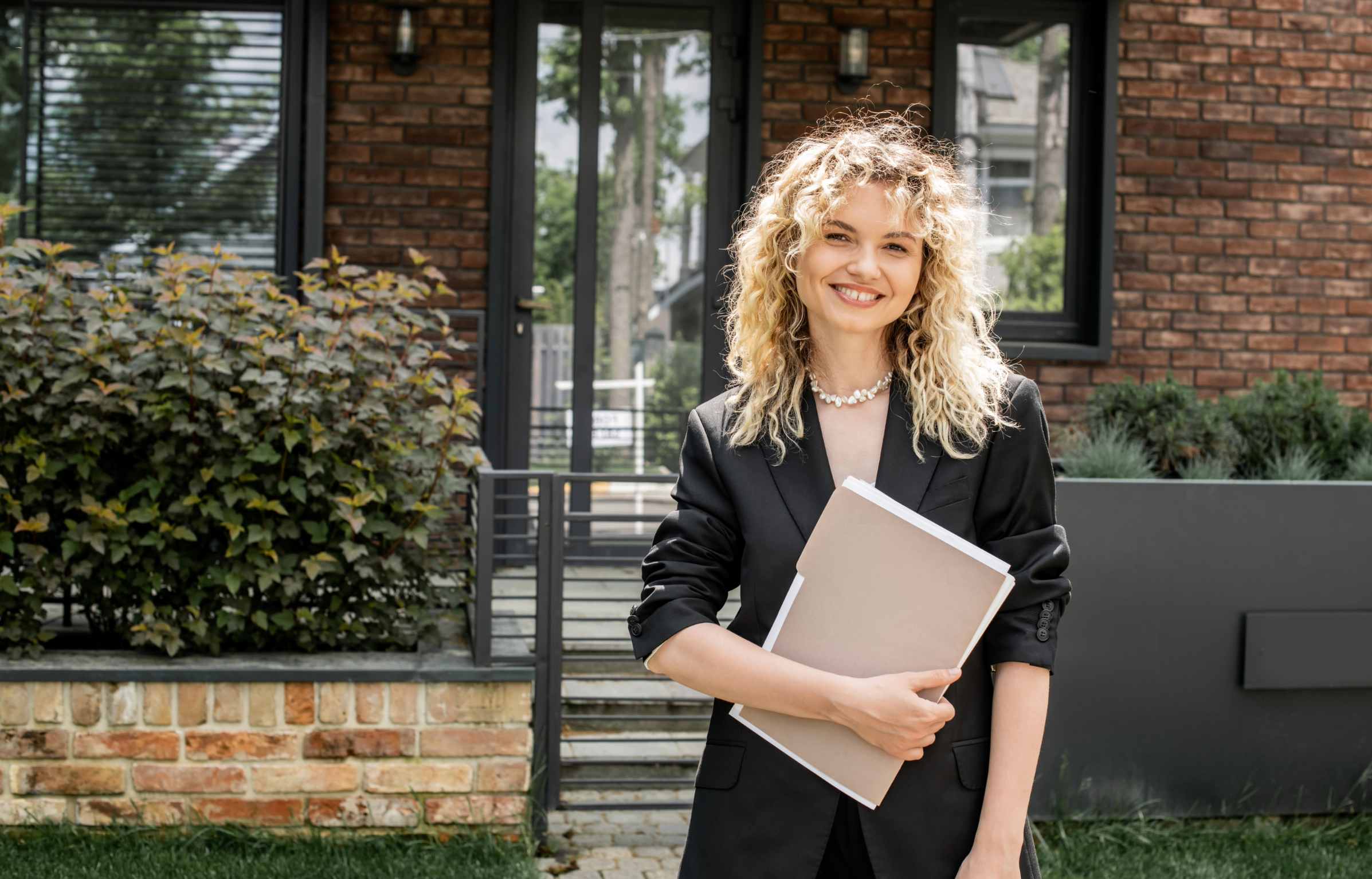 cheerful blonde real estate agent holding documents and looking at camera near house on city street