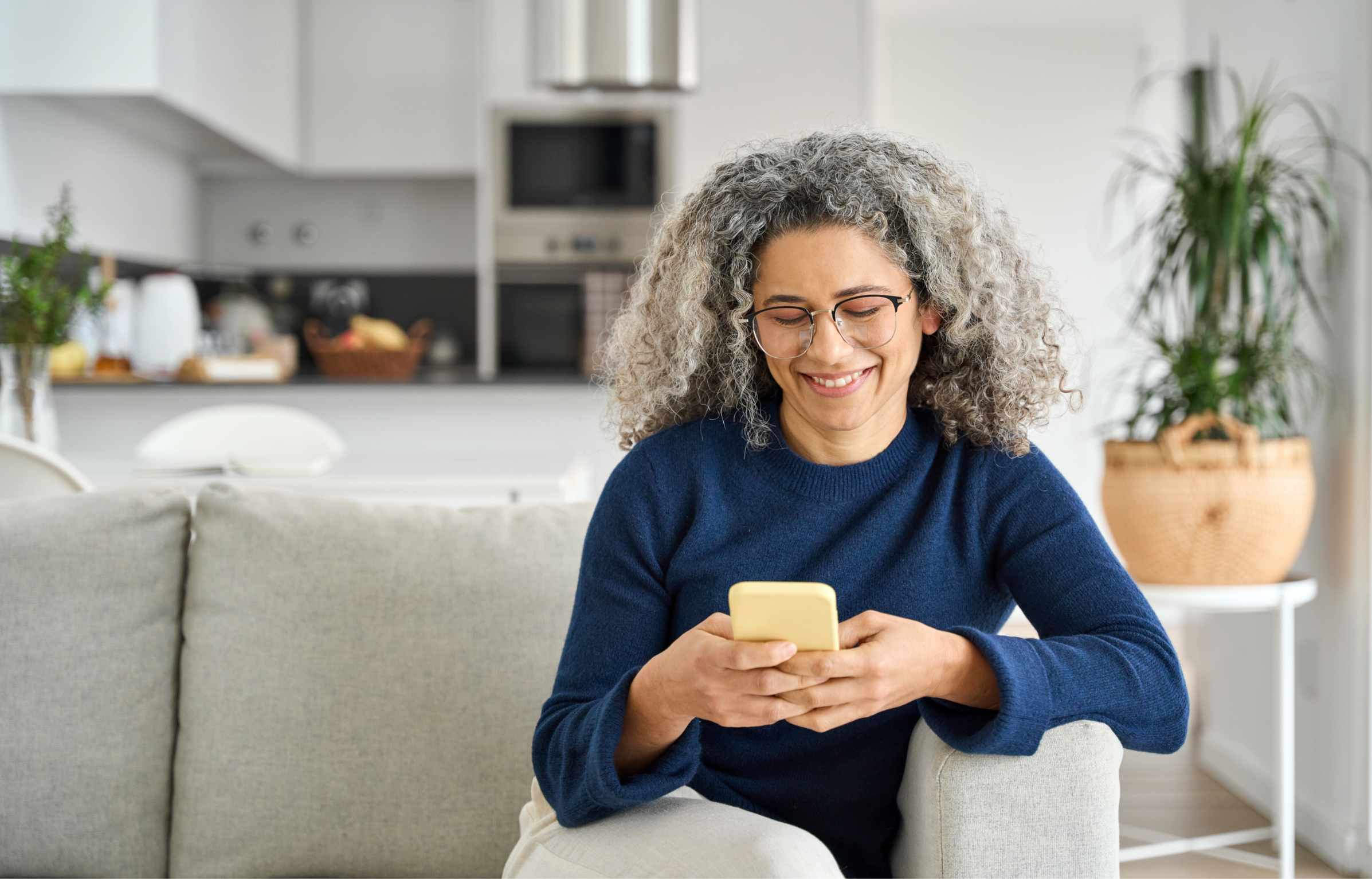 Happy middle aged woman using smartphone sitting on couch at home. Mature lady wearing glasses holding cellphone browsing internet, texting messages on mobile cell phone technology on sofa