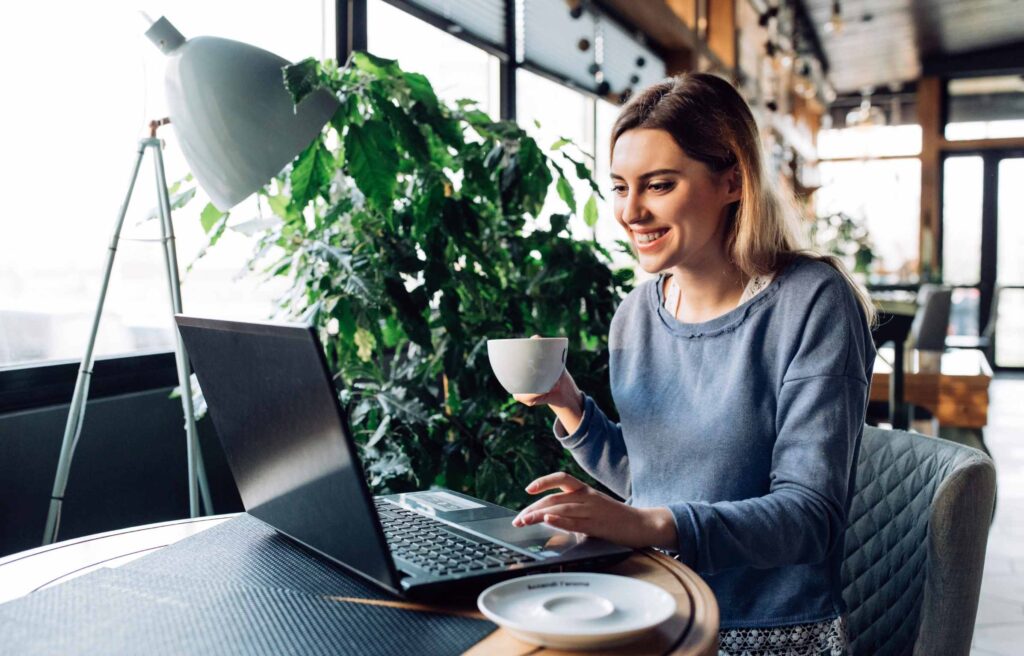 Cheerful pretty girl looking at laptop screen, holds a cup of coffee, while resting at cafe.