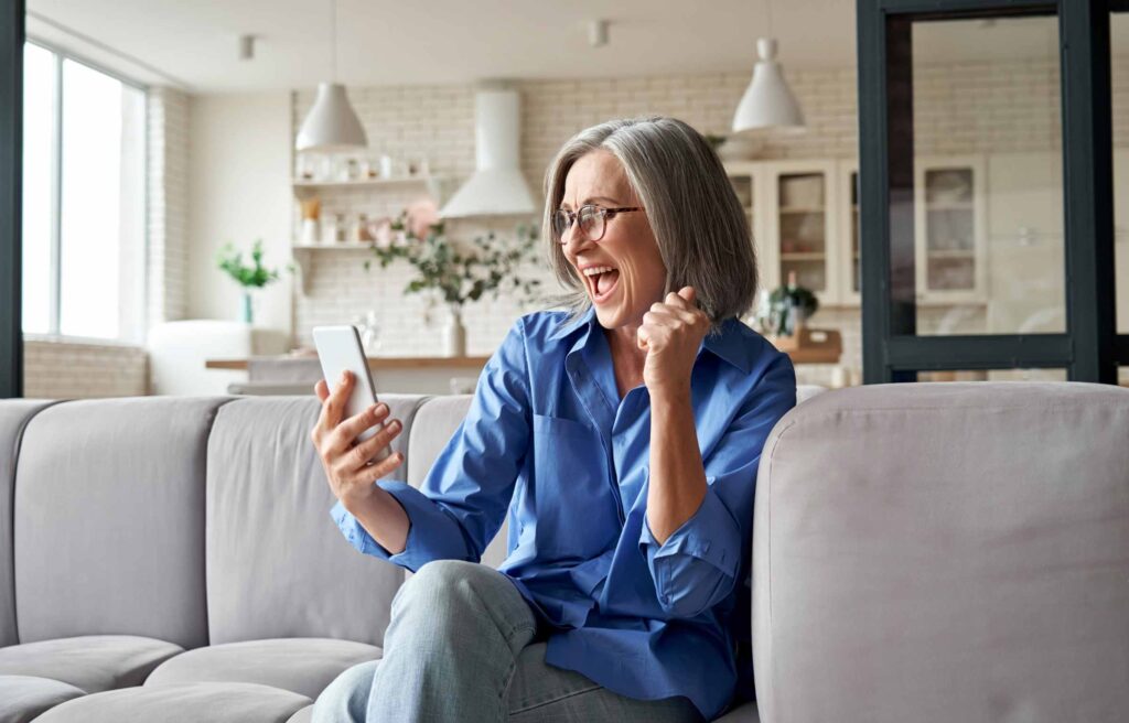 Excited happy mature old 60s woman customer winner holding smartphone using mobile app winning online lottery bid, celebrating success, receiving gift voucher on cell phone sitting on couch at home