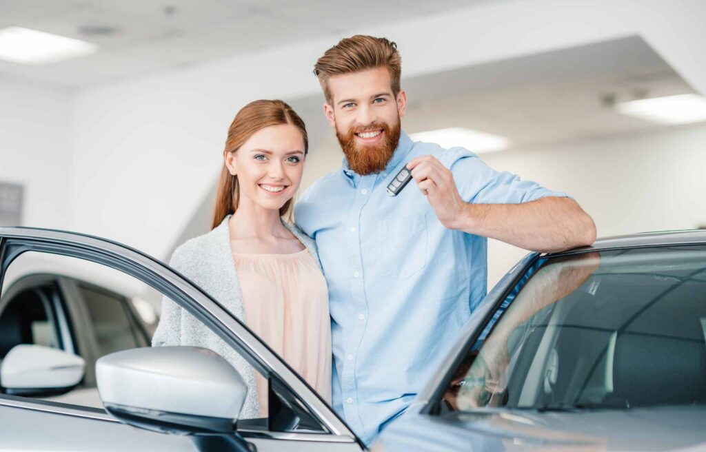 Happy couple holding car key and standing at car in dealership salon