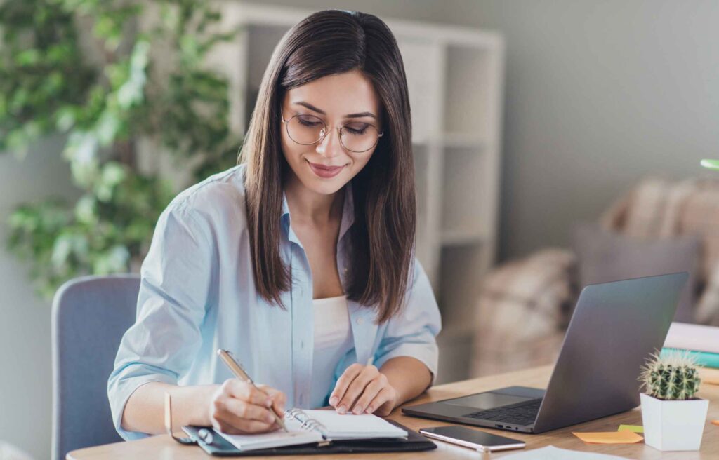 Photo of pretty young girl sit desktop pc hold pen write notepad wear glasses shirt in home office indoors