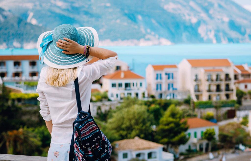 Tourist woman admiring view of colorful tranquil village Assos on morning. Young stylish female model wearing blue sunhat and white clothes enjoying summer travel vacation in Kefalonia, Greece