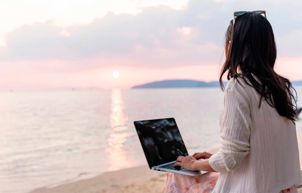 Young woman freelancer traveler working online using a laptop and enjoying the beautiful nature with sea view at sunset