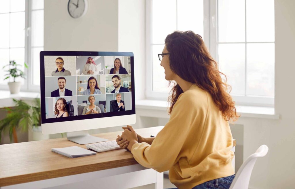 Corporate business team having a remote meeting. Young woman in casual clothes sitting at her desk at home, attending an online conference, looking at the computer screen, and smiling