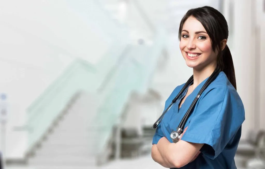 Smiling nurse standing with arms crossed, wearing blue scrubs and a stethoscope in a clinical setting, exploring relocation packages for nurses.