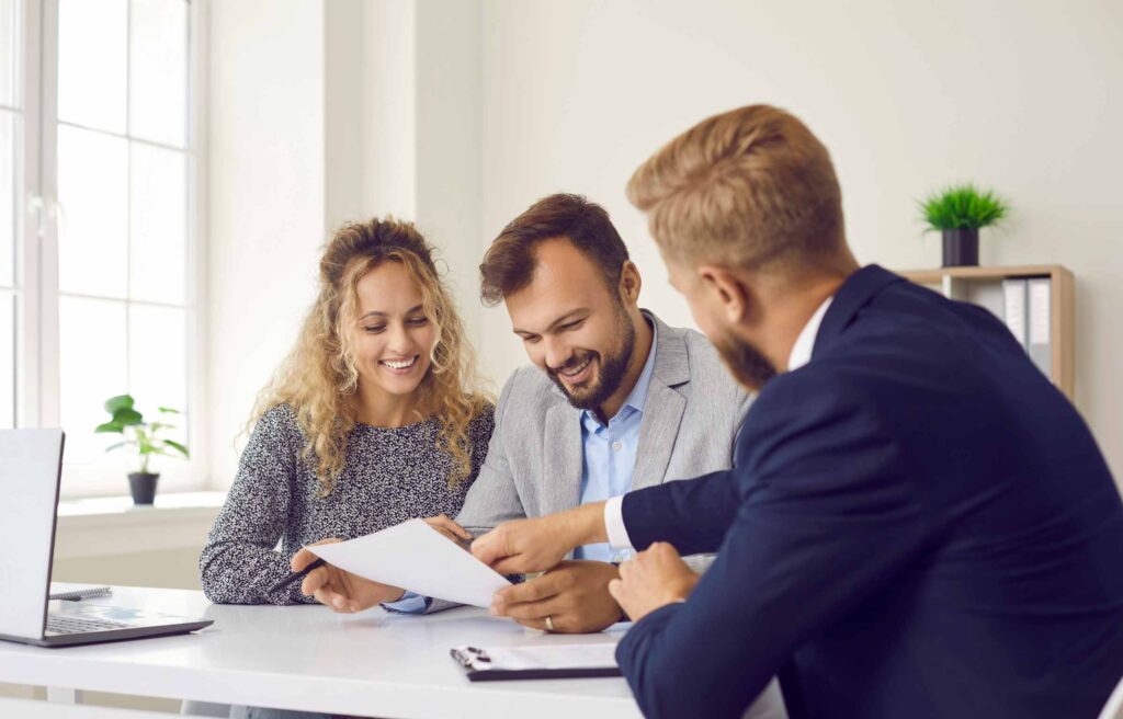 Happy young family at meeting in office signs document on purchase of real estate or loan agreement. Head of bank, realtor or financial advisor submits contract and indicates place of its signing