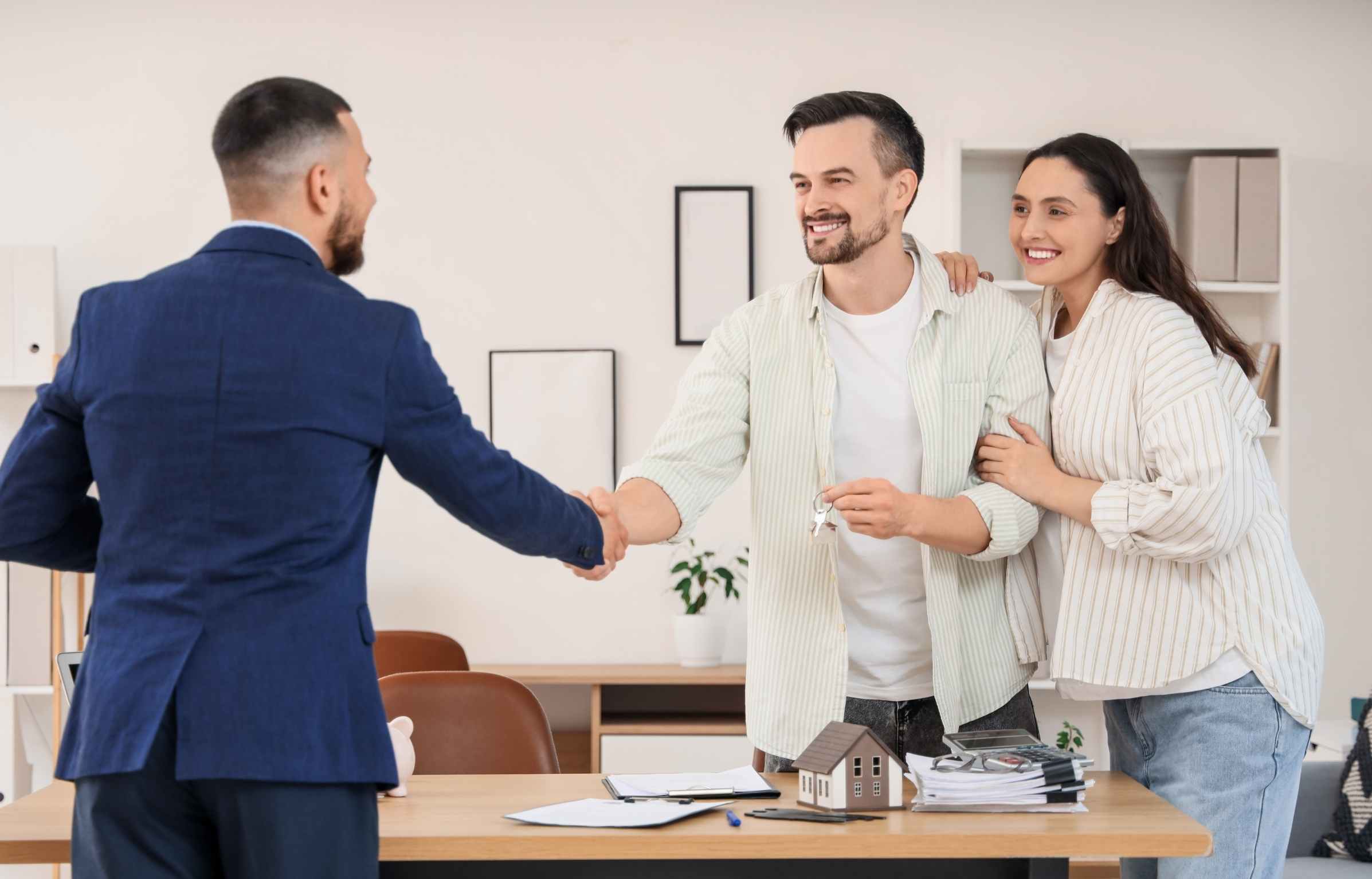 Male Real Estate Agent and Young Pregnant Couple with Key from House Shaking Hands in Office