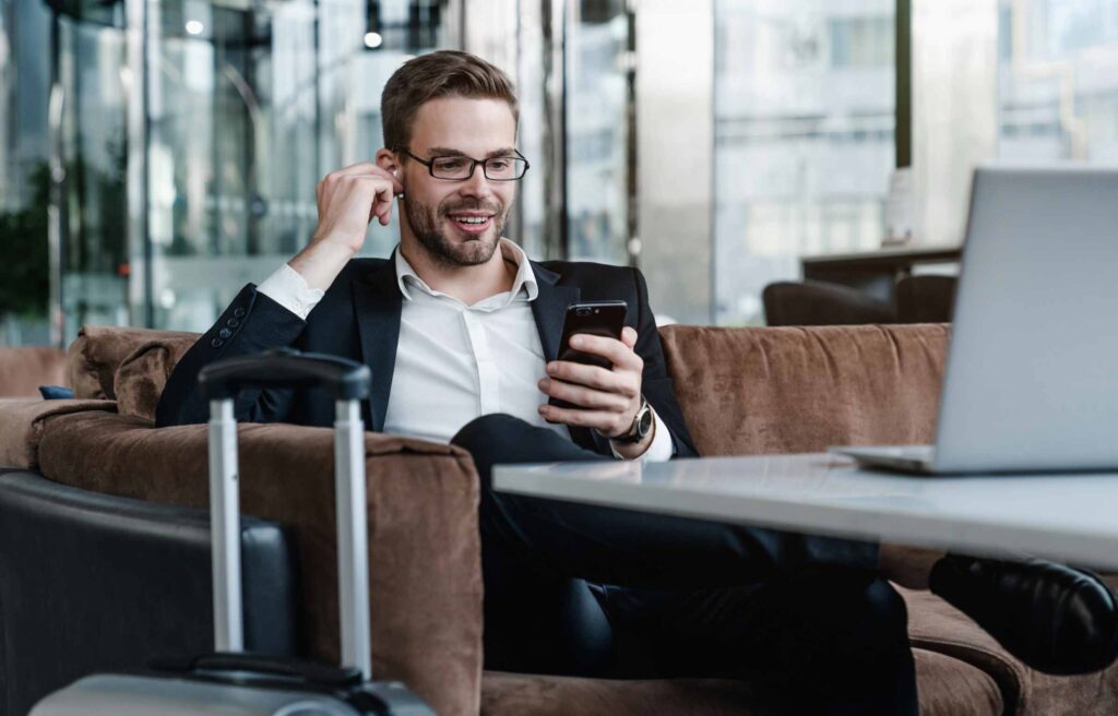 Man with suitcase sitting in airport waiting area while listening music using airpods