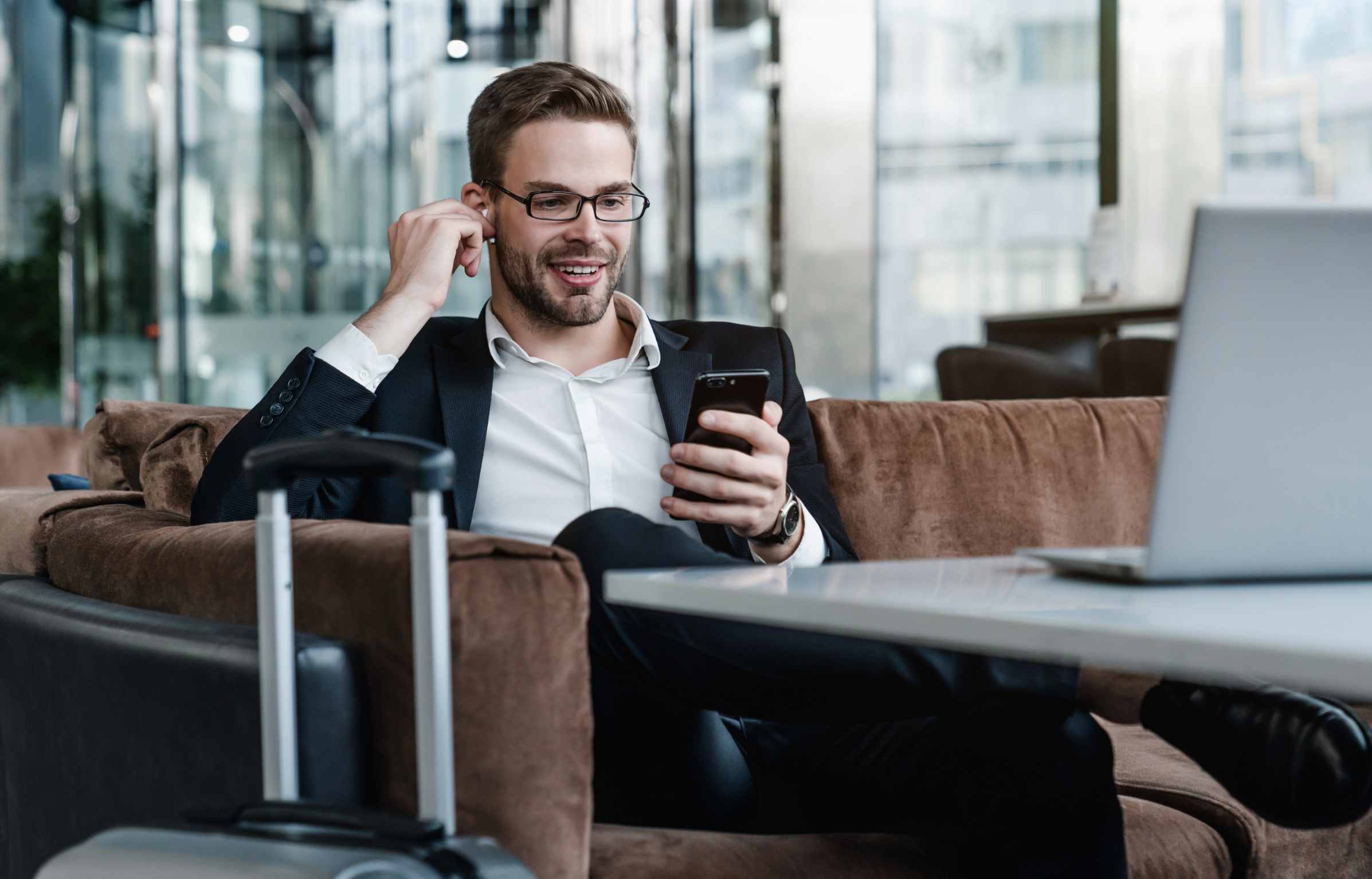 Man with suitcase sitting in airport waiting area while listening music using airpods
