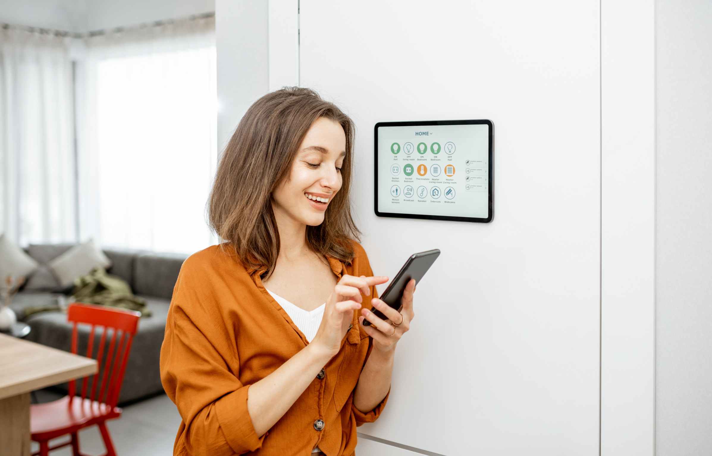 Young woman controlling home with a digital touch screen panel installed on the wall in the living room. Concept of a smart home and mobile application for managing smart devices at home