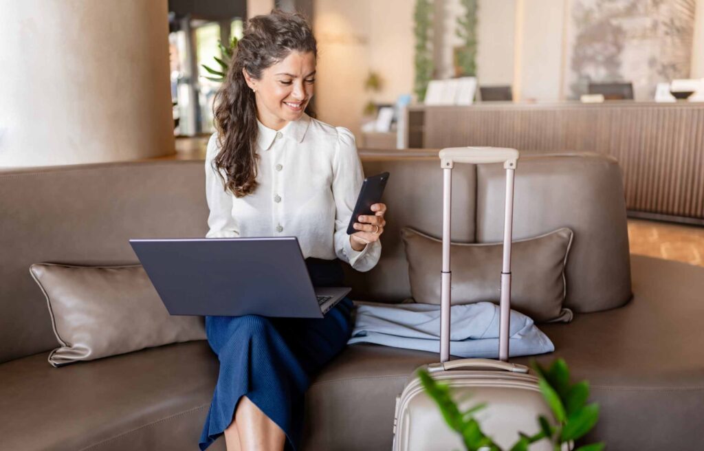 A woman sits in a hotel lobby using a laptop while her suitcase stands nearby. She appears focused on her work in a comfortable, modern capital one lounge access changes area.