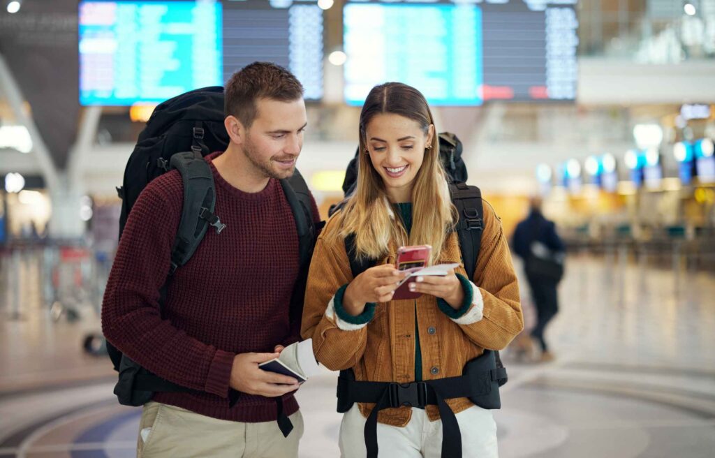 Happy couple, airport and phone with ticket, travel app and adventure with excited face, conversation and smile. Man, woman and smartphone for digital booking of hotel, taxi or bus for transportat