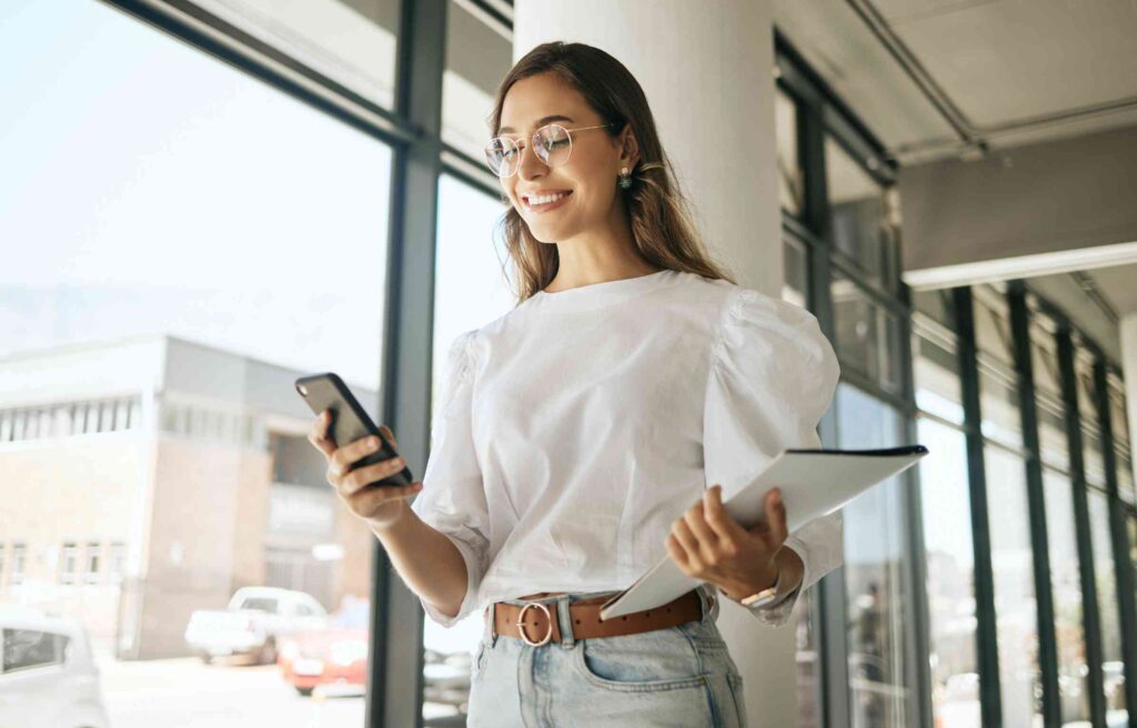 A woman employee checks employee relocation services on her phone while holding a tablet near a bright office window.