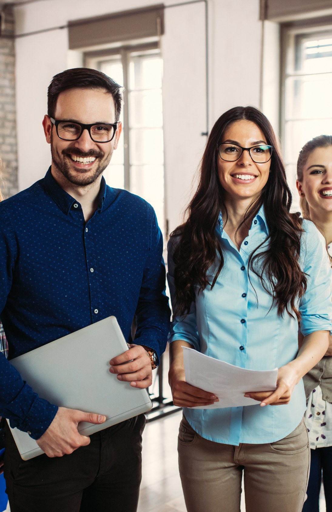 Two smiling corporate employees wearing glasses, standing together in an office environment, holding documents and a laptop.
