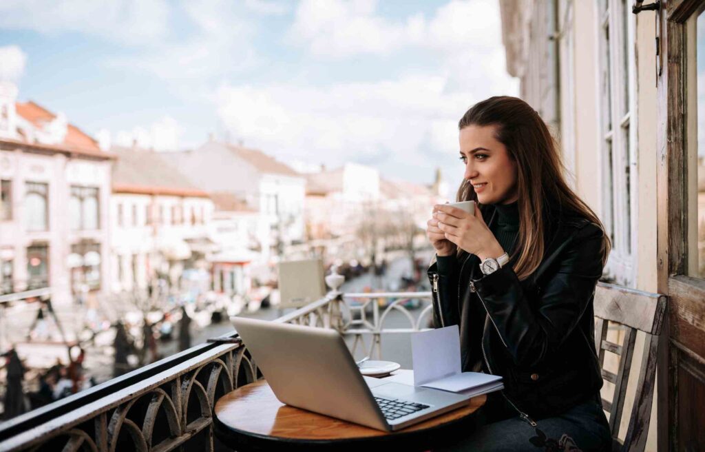 Digital nomad woman in a European city sitting on a balcony with a laptop and coffee, overlooking a scenic street view.