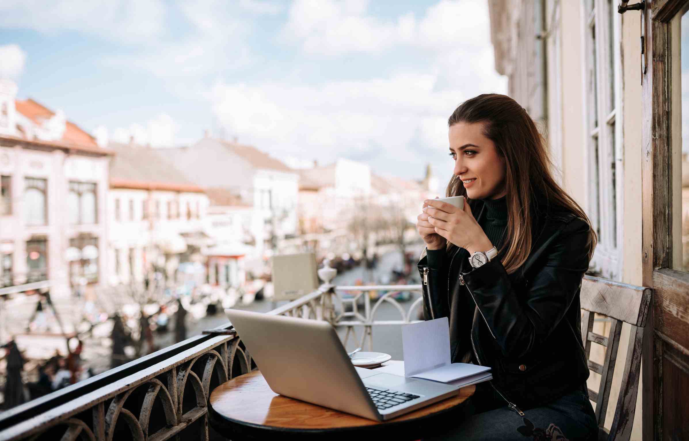 Digital nomad woman in a European city sitting on a balcony with a laptop and coffee, overlooking a scenic street view.