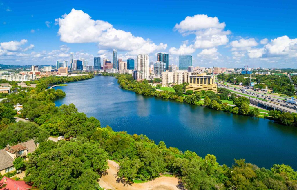 Aerial view of downtown Austin, Texas with skyscrapers, the Colorado River, and green parks under a bright blue sky.