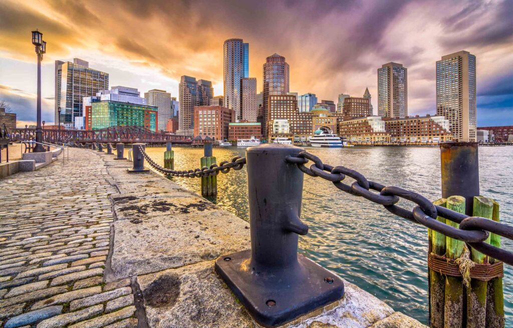 Boston waterfront with cobblestone path, harbor, and skyline of modern high-rise buildings at sunset.