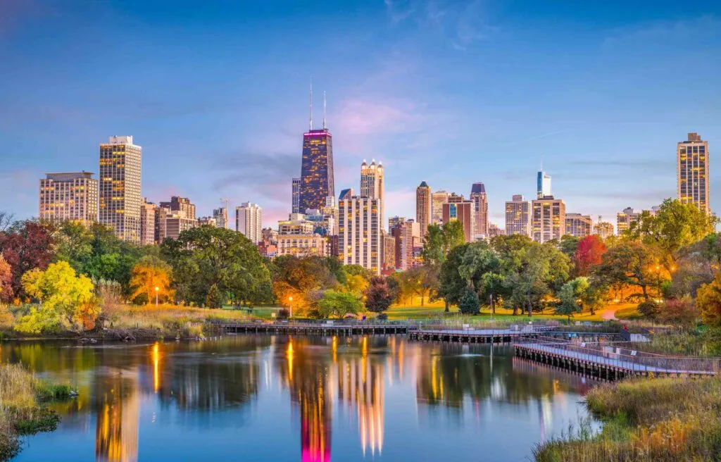 Chicago Illinois skyline at twilight with city buildings reflected in a calm lake and trees in the foreground.
