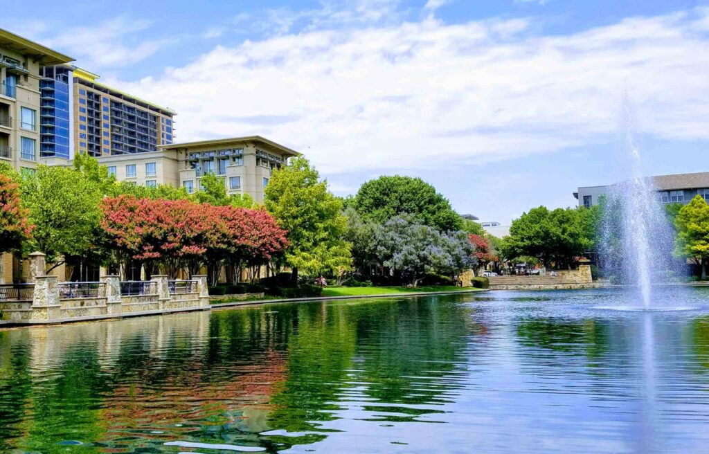 Scenic view of a lake with a fountain, surrounded by modern buildings, green trees, and colorful landscaping in Plano, Texas.