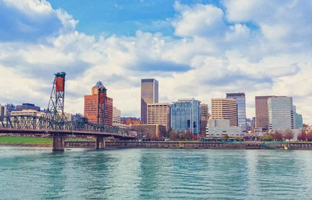 Portland Oregon city skyline with bridge crossing the Willamette River under a partly cloudy sky.
