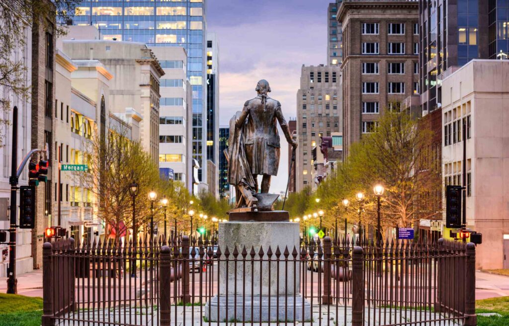 Downtown Raleigh, North Carolina, with a statue in the foreground and modern high-rise buildings in the background at dusk.