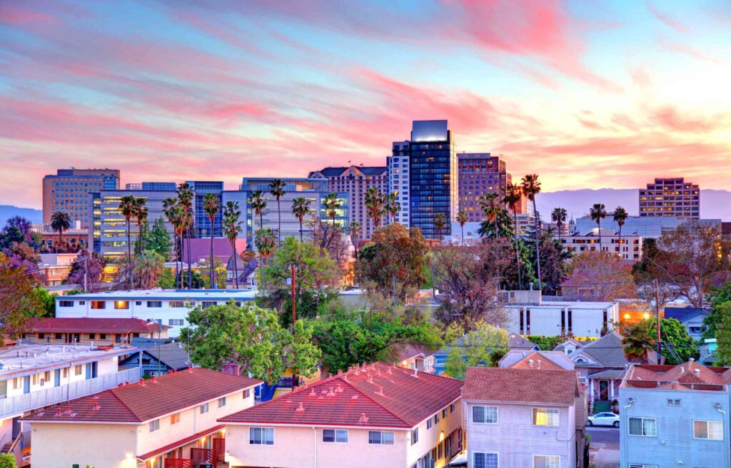 San Jose skyline at sunset with palm trees, modern office buildings, and residential rooftops in the foreground.