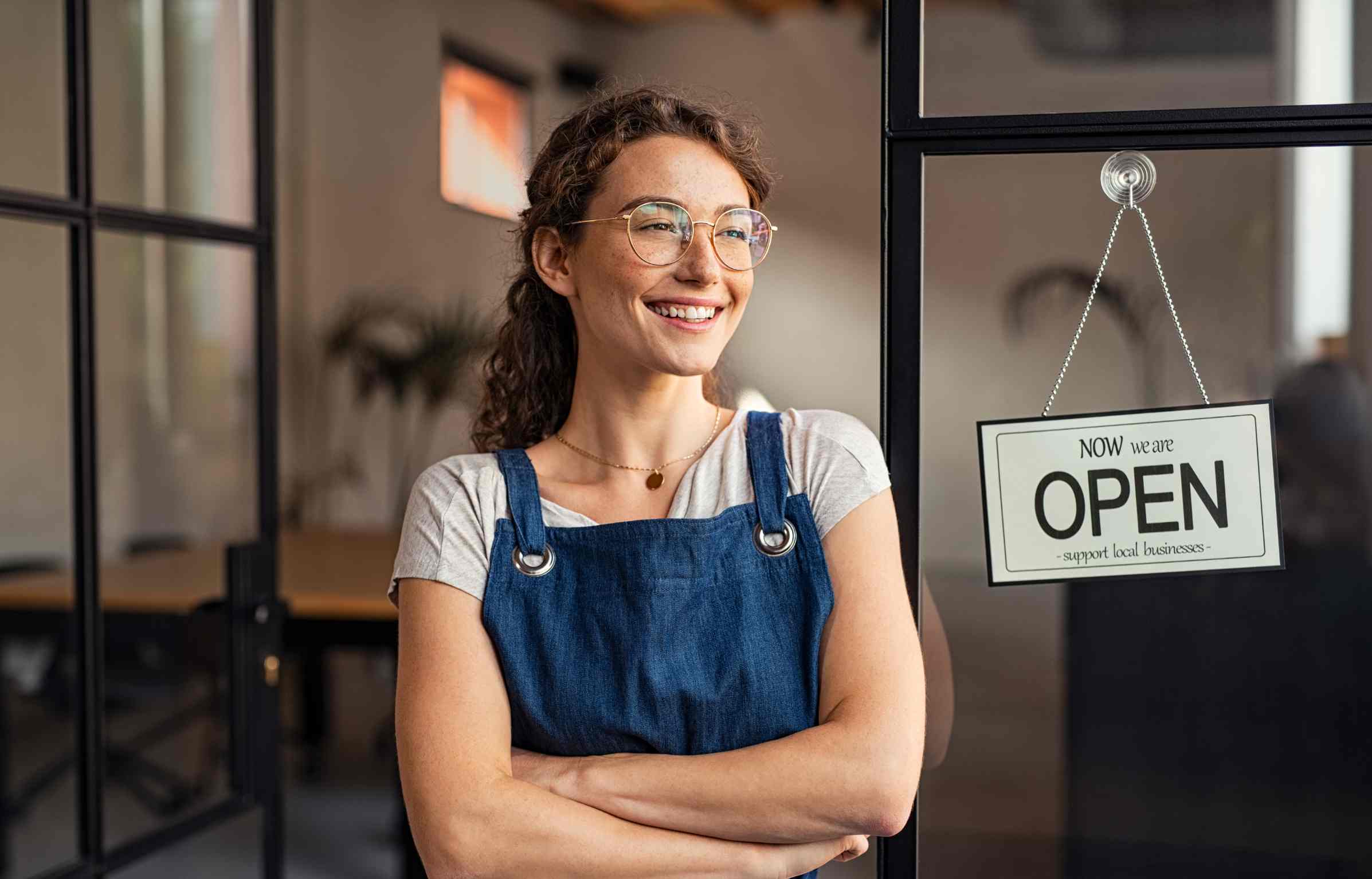 Smiling small business owner at shop entrance with open sign, showing how free rewards programs and daily cashback support growth.