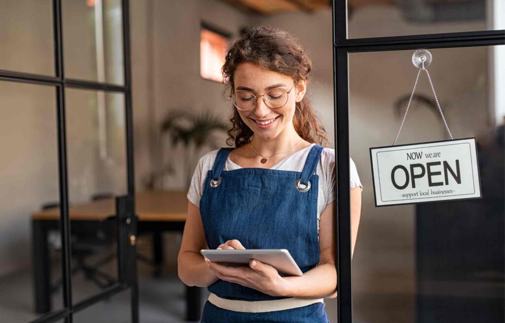 Small business owner standing at shop entrance with open sign, smiling while checking rewards on a tablet, showing the benefits of a free rewards program for small business.