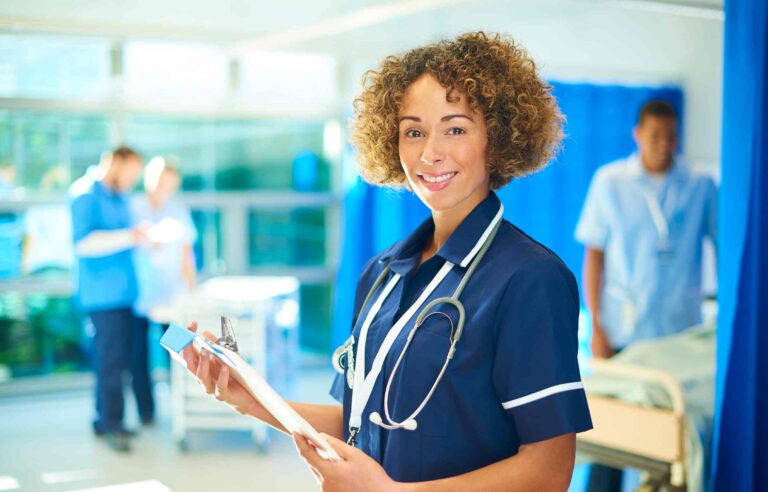 Smiling registered nurse in blue uniform with clipboard and stethoscope in a hospital ward.