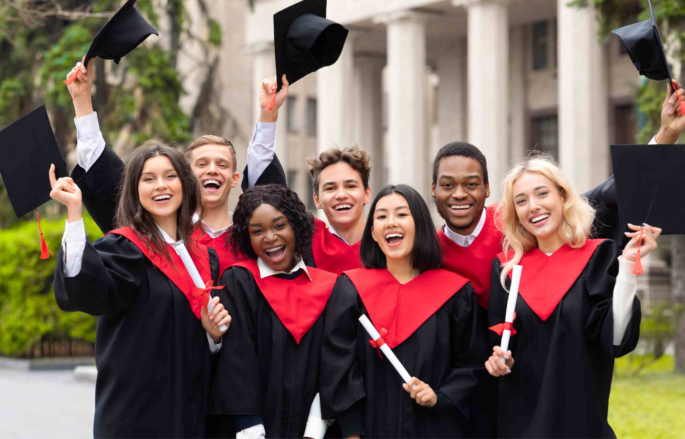 Group of diverse graduates in caps and gowns celebrating with diplomas and raised hats outside a university building.