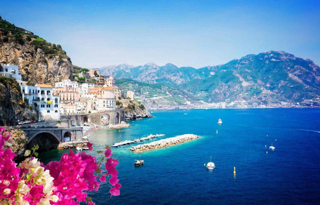 Colorful houses built on cliffs overlooking the blue waters of the Amalfi Coast in Italy with pink flowers in the foreground and mountains in the background.